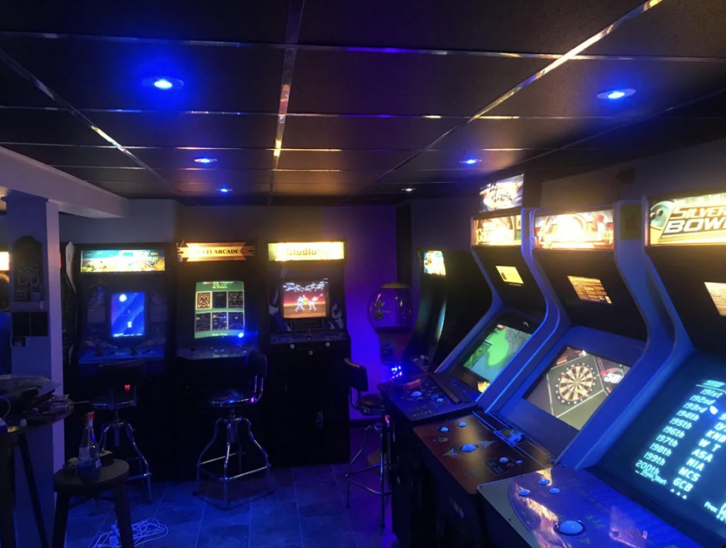 Dimly lit arcade room featuring multiple classic arcade game machines with bright screens and illuminated marquees. The room has a futuristic ambiance with blue neon lights on the ceiling. A few bar stools are placed in front of the machines.