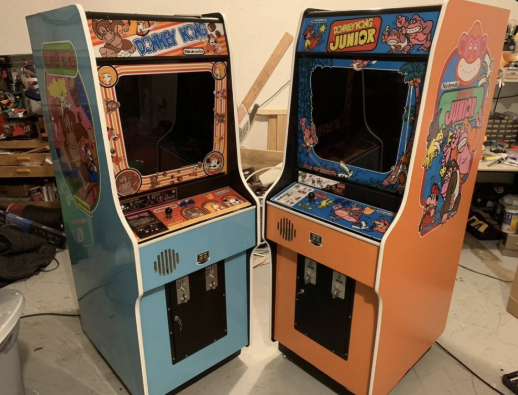 Two vintage arcade machines are displayed. The left machine, themed in light blue, features the game "Donkey Kong," while the right machine, themed in orange, displays "Donkey Kong Junior." Both machines showcase colorful artwork and classic joystick controls.