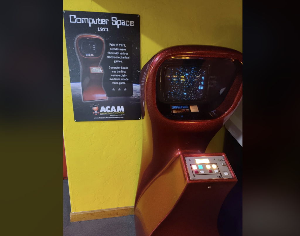 A vintage "Computer Space" arcade cabinet from 1971 is displayed against a yellow wall. Next to it is a poster explaining the historical significance of the game. The arcade machine has a curvy, red metallic exterior and a control panel with buttons and a joystick.