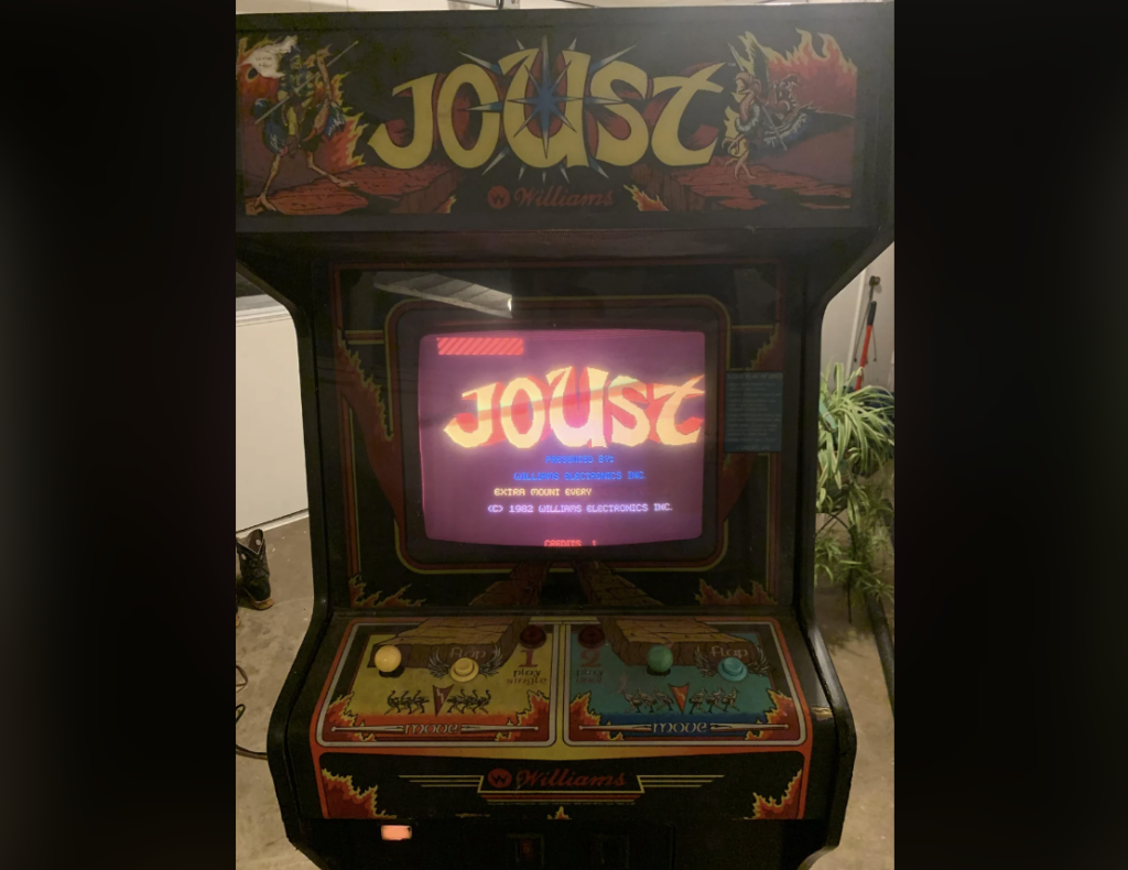 A classic Joust arcade game machine is prominently displayed, featuring vibrant artwork and a screen showing the game's title. The control panel includes joysticks and buttons for two players, adorned with game-themed decals and instructions.