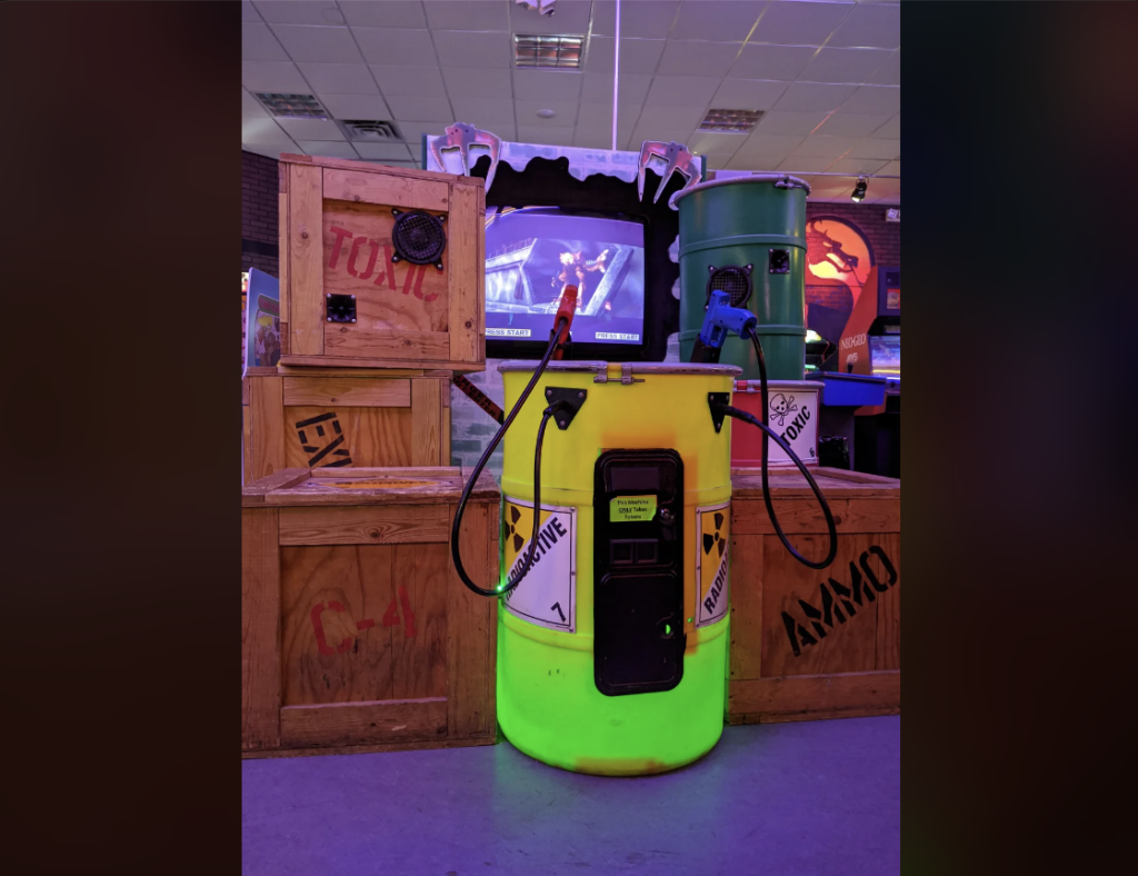 A neon green barrel with caution tape and a red axe leaning against it is surrounded by wooden crates. A green metal barrel and various props are in the background, along with a dimly lit arcade game screen. The atmosphere appears to be themed and immersive.