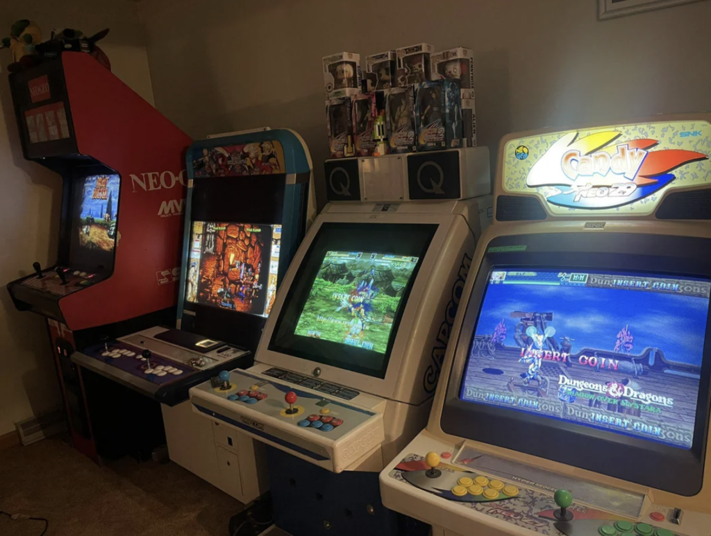 A room with several retro arcade cabinets lined up. From left to right, there's a Neo Geo MVS, a machine with Marvel vs. Capcom, a Capcom arcade cabinet featuring Darkstalkers, and another Capcom cabinet displaying Dungeons & Dragons. Figures sit atop two cabinets.