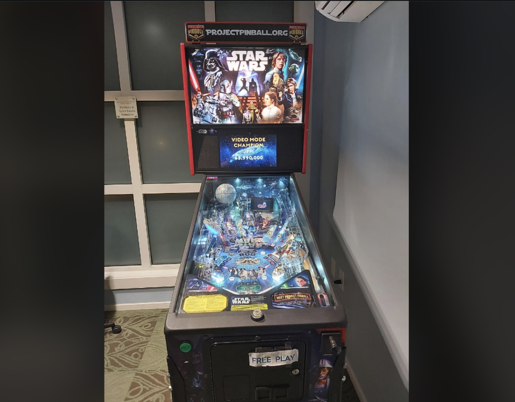 An angled view of a Star Wars-themed pinball machine situated in a corner of a room. The machine features vibrant artwork of Star Wars characters and is on free play. The back panel displays "PROJECTPINBALL.ORG" and a high score, while ambient lighting illuminates the playfield.