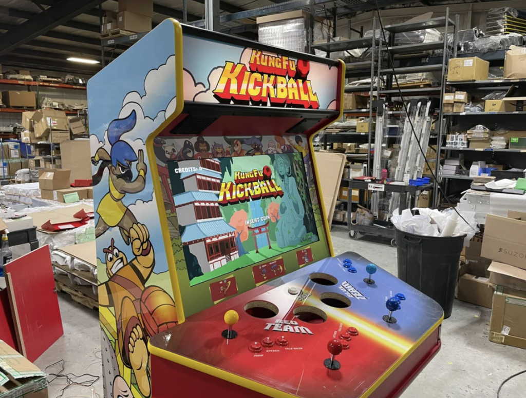 An arcade machine featuring the game "Kung Fu Kickball" stands in a cluttered warehouse. The machine is colorful with characters and game imagery on the sides and front. Surrounding the arcade machine are various boxes, shelves, and scattered items.