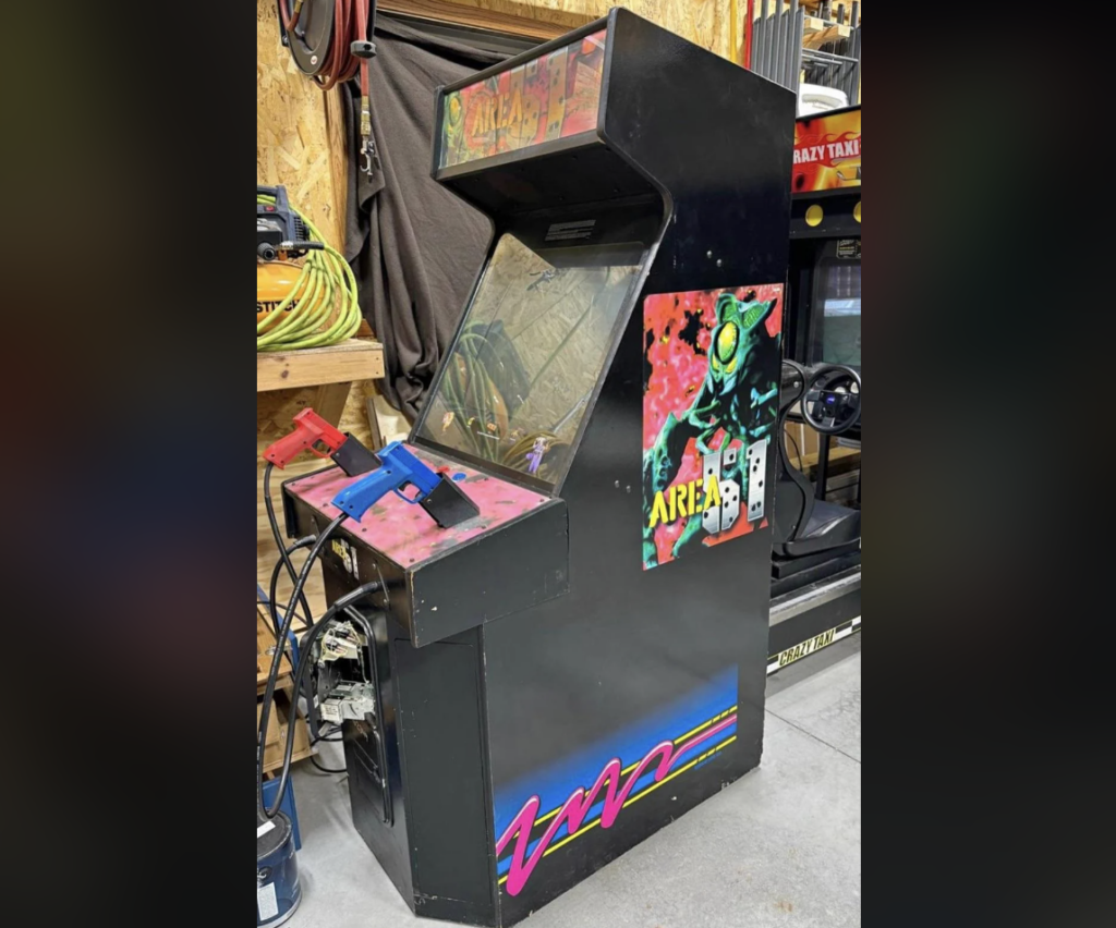 An upright arcade cabinet with the game "Area 51" displayed. The cabinet is black with vibrant, colorful graphics, including a futuristic soldier on the side. Attached to the front panel are blue and red light guns. The arcade cabinet is placed in a garage or workshop setting.