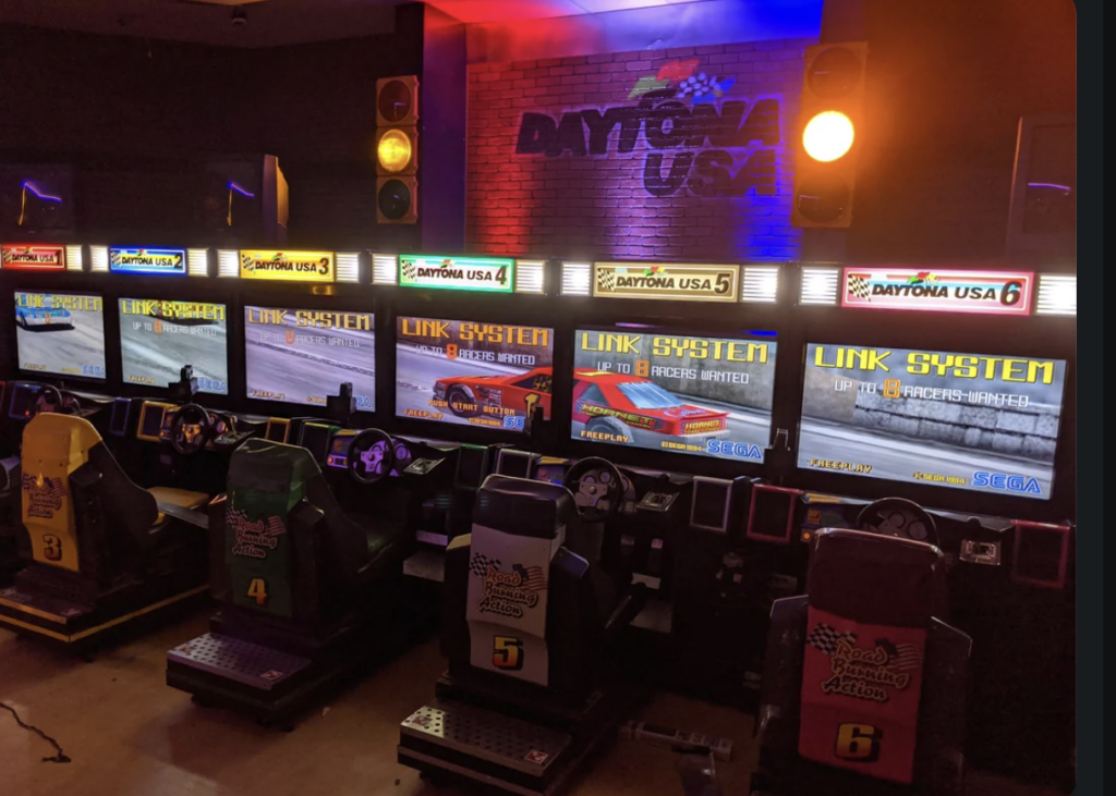 A row of six connected arcade racing game machines with the title "Daytona USA" above them. Each machine has a steering wheel, pedals, and a racing seat. The screens display the game's interface with a nighttime gaming arcade ambiance in the background.