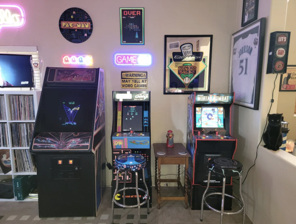 A room filled with vintage arcade games, including Pac-Man, Mortal Kombat, and others. Neon signs and framed memorabilia adorn the walls. A black cat sits on a table next to the games. Vinyl records are partially visible on shelving to the left.