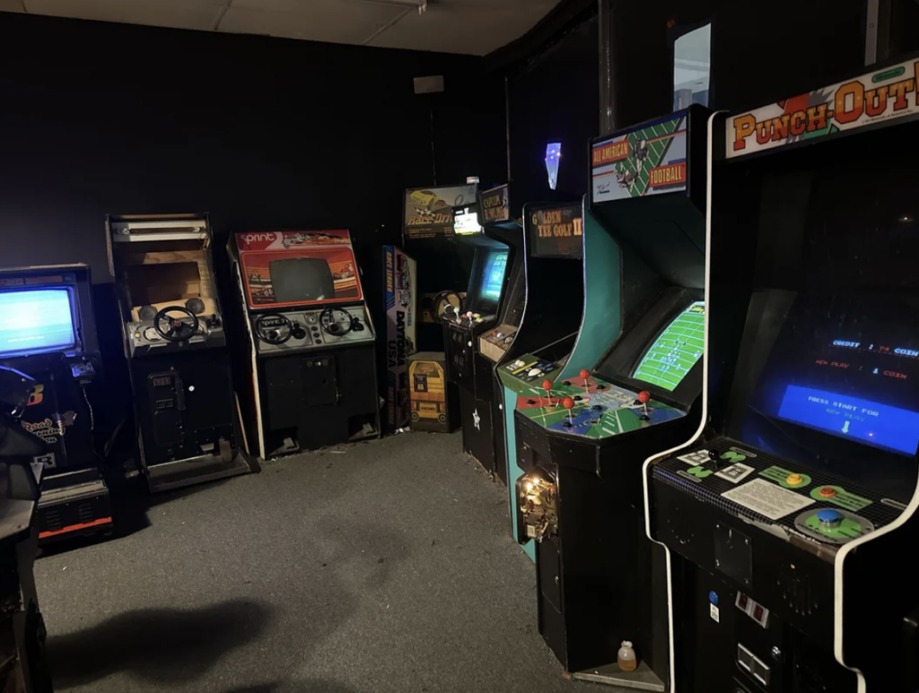 A dimly lit arcade room filled with vintage arcade machines. Titles include "Punch-Out!!" and various racing and shooting games. The carpeted floor and dark walls add to the retro atmosphere. Some screens are illuminated with games in progress.