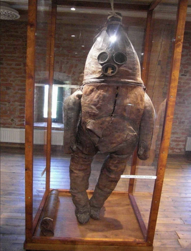 A deteriorating antique diving suit, resembling a large bulbous body with a cylindrical helmet and round glass portholes, displayed in a wooden and glass showcase within a room with exposed brick walls and wooden flooring. A label is visible near the suit's feet.