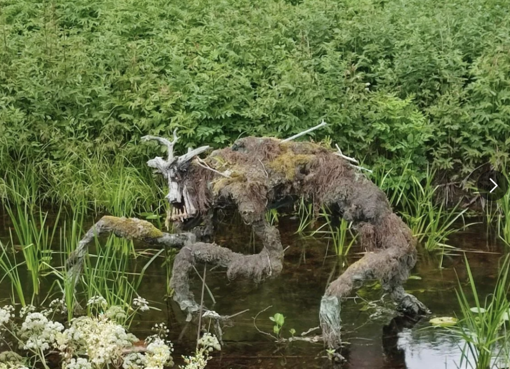 A sculpture resembling a creepy, moss-covered creature with elongated limbs and a gnarled face emerges from a marshy area with dense greenery in the background. The creature has long, twisted arms and appears to be moving through the shallow water.