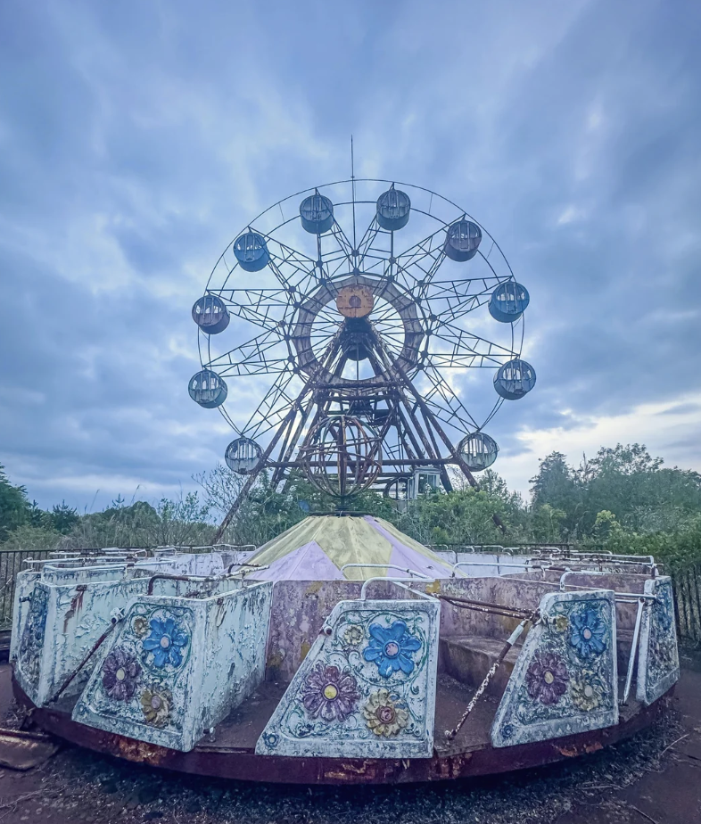 An old, abandoned amusement park ride sits in the foreground, covered in chipped paint with floral designs. In the background, a large, rusting Ferris wheel looms against a cloudy sky, adding to the eerie atmosphere of the scene.