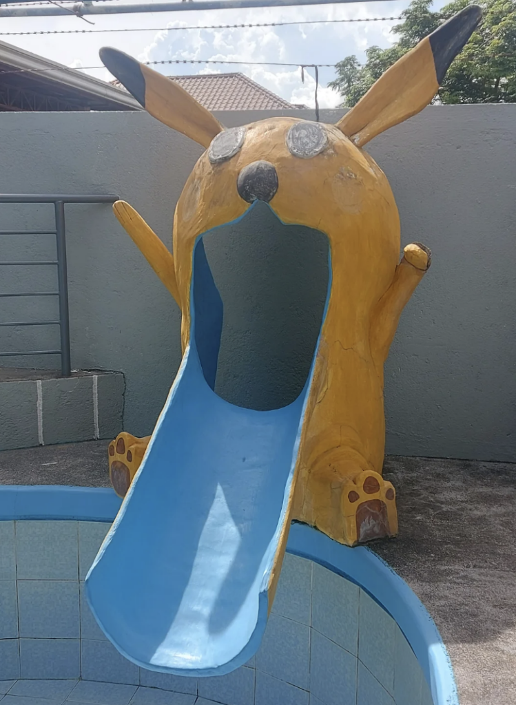 A brightly painted yellow slide shaped like a character with ears and paws, featuring a wide open mouth serving as the slide entrance. The slide itself is blue and leads down into a shallow tiled pool area. The background includes a grey wall and a railing.