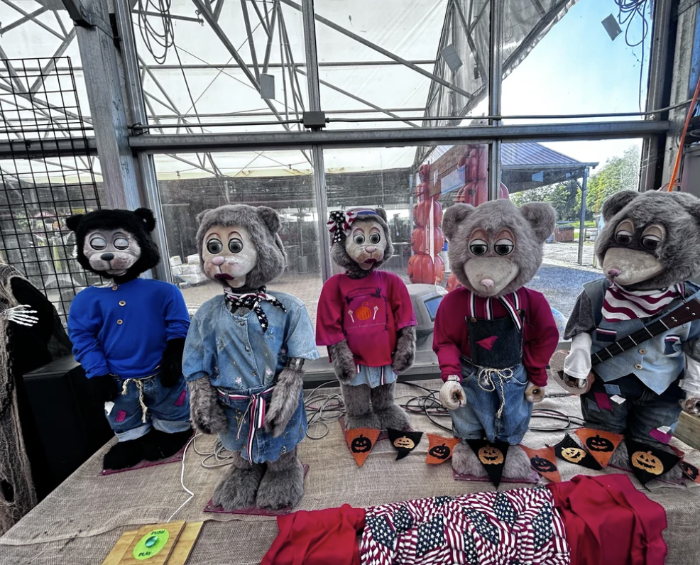 A group of five anthropomorphic bear-like figures, dressed in varied and colorful outfits, are arranged in a row on a table. One wears a black hat and another has a pink bow. Pumpkin-themed bunting decorates the front of the table. They are displayed indoors.