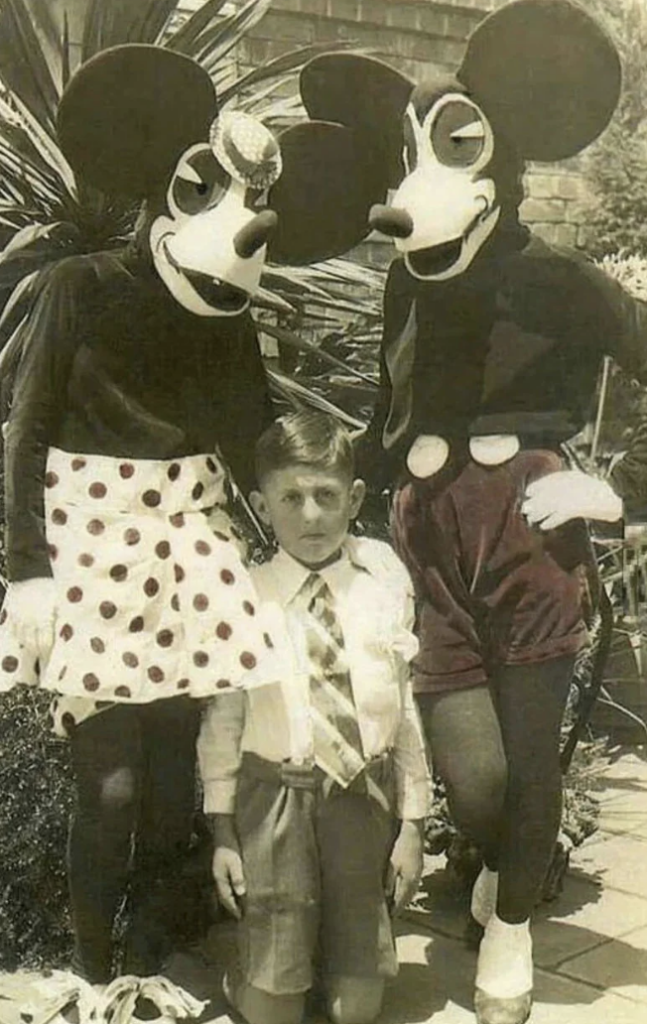 A black-and-white photo shows a young boy standing between two people dressed in old-fashioned Mickey and Minnie Mouse costumes, with exaggerated facial expressions. The boy appears serious while the costumed characters stand on either side of him. A plant is in the background.