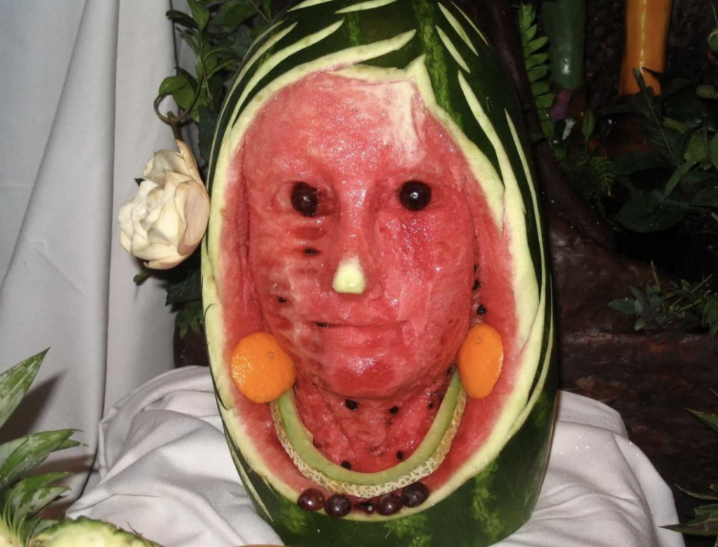 A carved watermelon depicted as a human face, adorned with fruit and vegetable decorations. The face uses watermelon flesh, with black seeds forming eyes and mouth details. Other fruits and vegetables create additional features, set on a white cloth background.