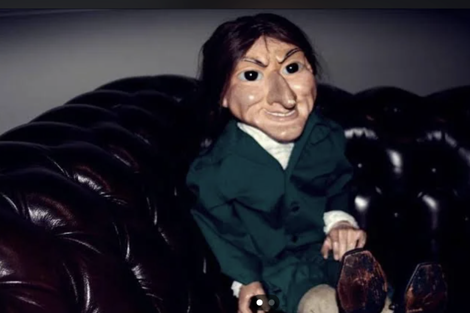 A puppet with exaggerated facial features, wearing a green outfit with white cuffs, is sitting on a dark leather couch. The background is dimly lit, making the puppet the clear focal point of the image.