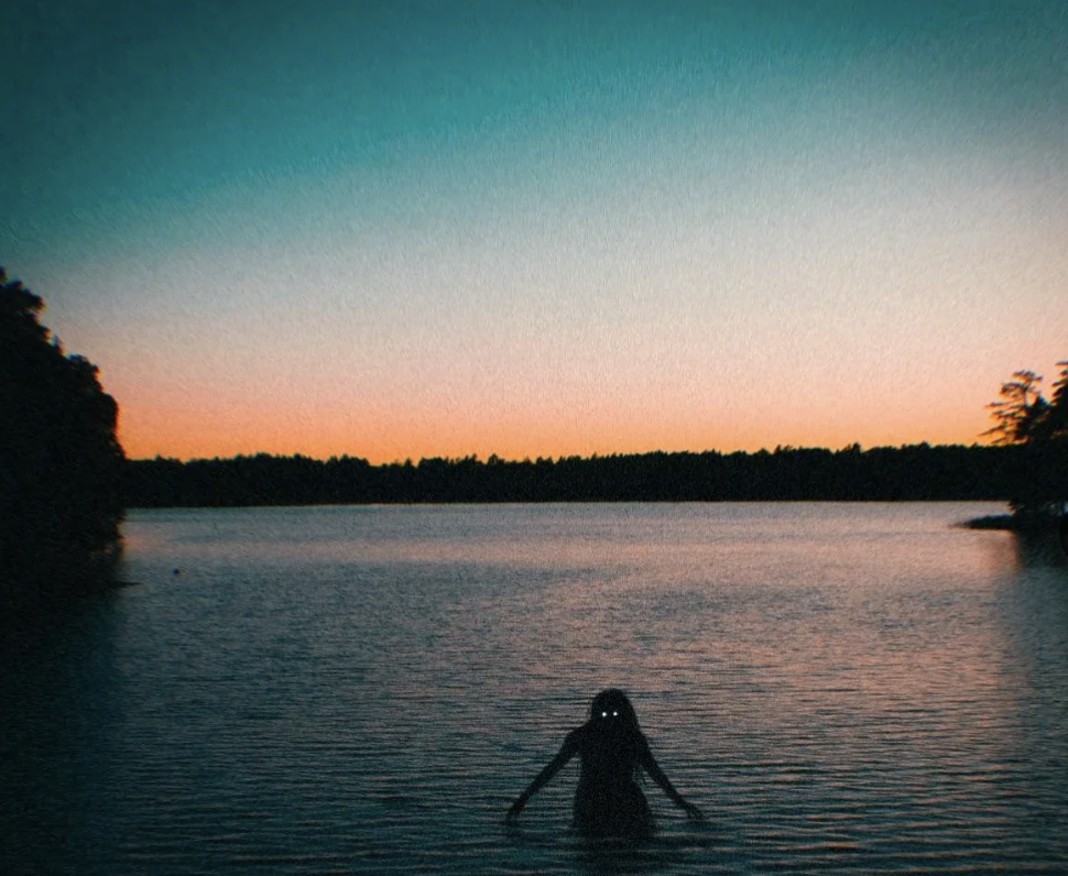 A silhouette of a person with glowing eyes stands in a calm body of water at dusk. The sky is transitioning from warm orange near the horizon to dark turquoise higher up. Trees line the horizon, and the water reflects the sky's gradient colors.