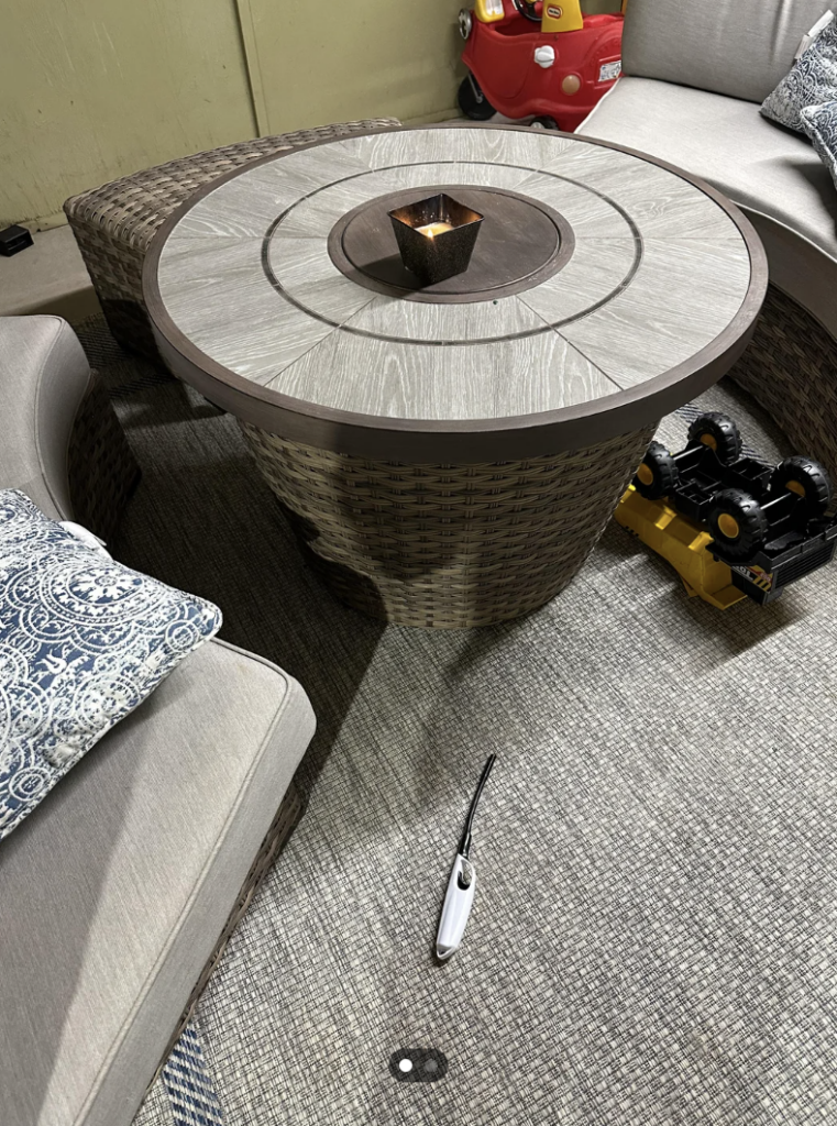 A circular outdoor table with a small candle at its center is surrounded by cushioned patio seating. Nearby, a toy fire truck and a lighter are on the floor. Decorative pillows with intricate patterns are on the seats. The area is carpeted with a textured rug.