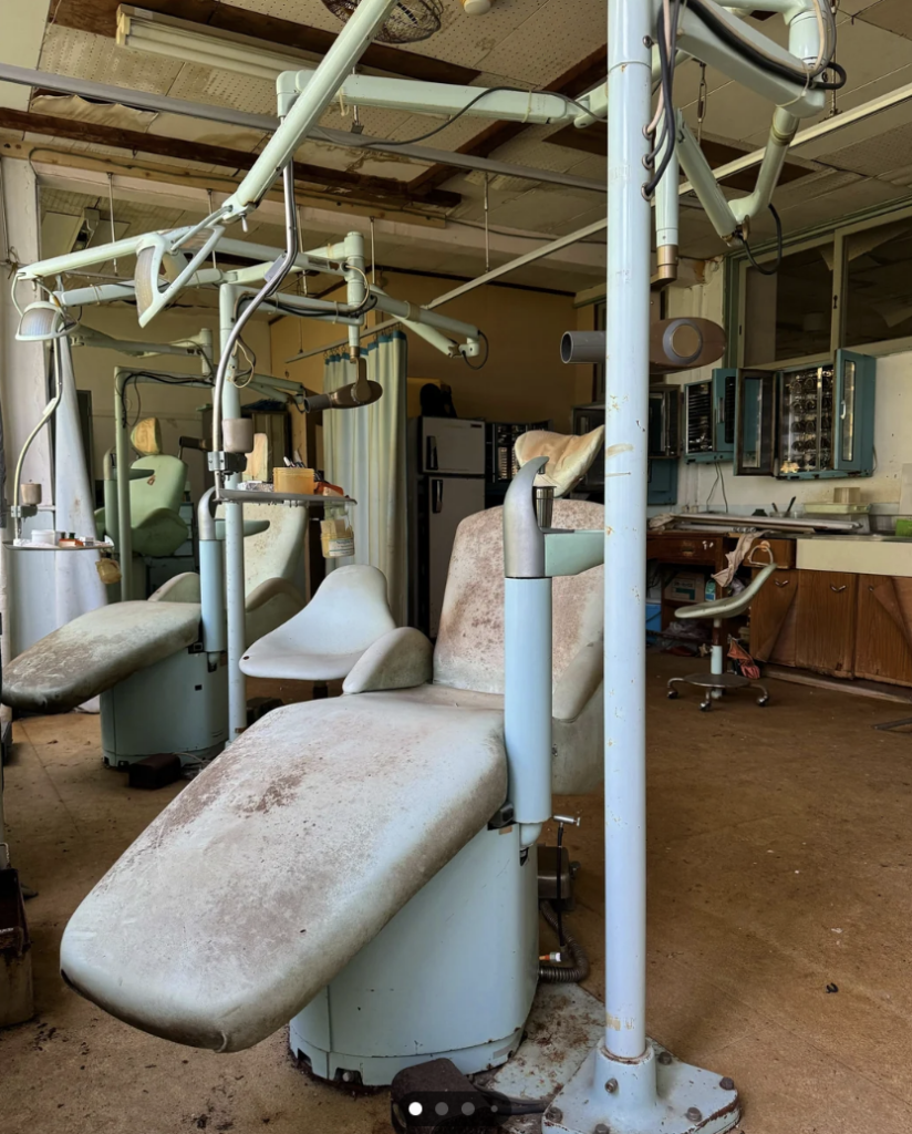 An abandoned dental clinic with rusty, dirty dental chairs and equipment. The ceiling appears damaged, and the room is cluttered with debris and old furniture. Everything looks outdated and neglected, conveying a sense of disuse and decay.