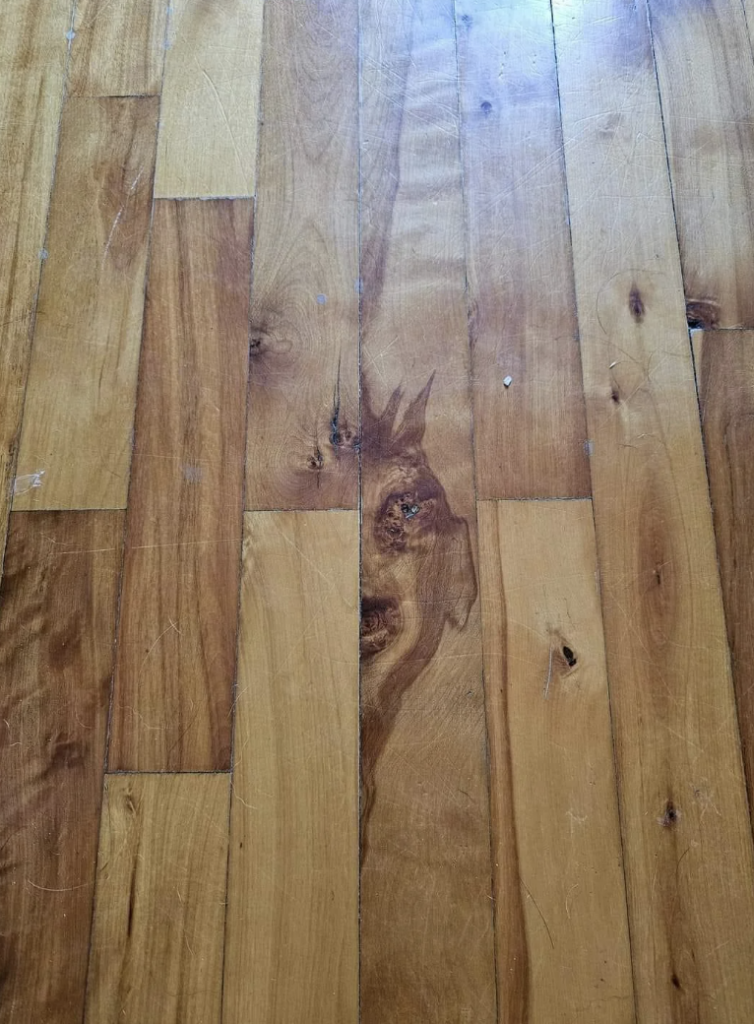 Wooden floorboards with a distinct natural pattern resembling a face towards the center. The face-like pattern includes two eyes, a nose, and a mouth formed by the wood grain and knots. The floor is light brown with some scratches and variations in color.