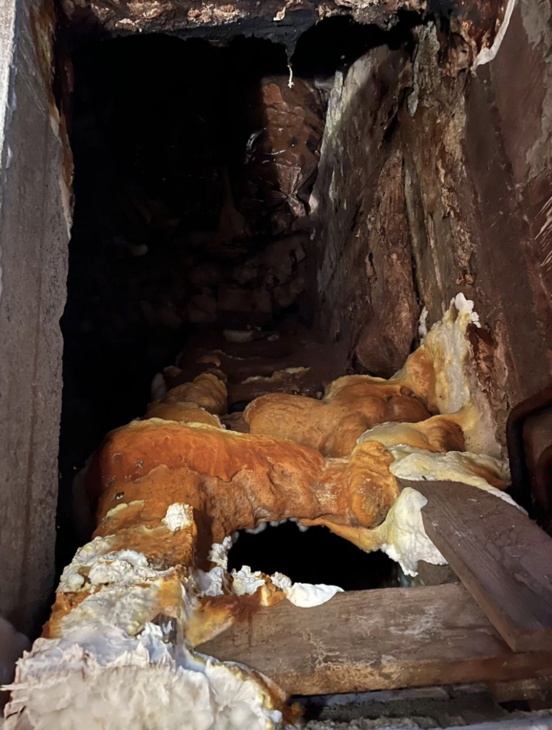 A narrow, vertical chimney space with dark, gritty walls partly illuminated, showing heavy buildup of thick layers of rust and mineral deposits. Wooden beams and various debris are lodged in the chimney structure. The scene has a neglected, decaying appearance.