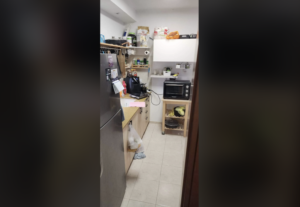 Compact kitchen with a silver refrigerator on the left, wooden cabinets and countertops, and various items on the shelves. The countertop holds kitchen appliances, dishes, and groceries. A toaster oven on a small cart is against the wall, with cabinets above.