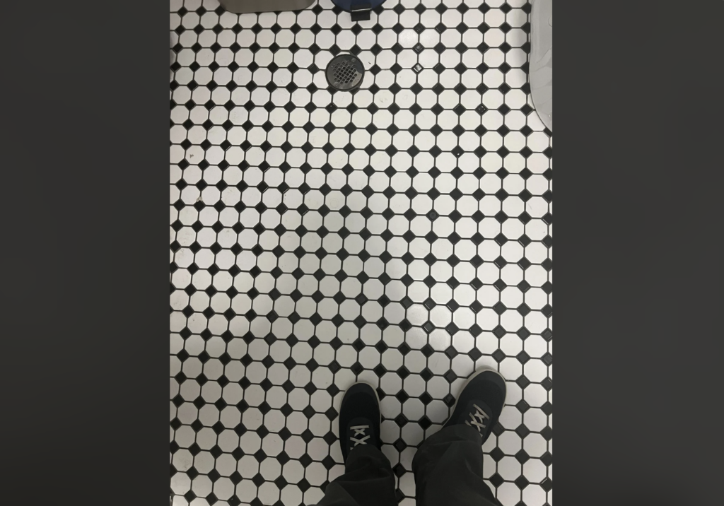 A person in black shoes is standing on a tile floor with a black and white hexagonal pattern. There's a circular drain in the center of the floor.