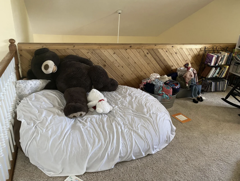 A cozy attic space with a large stuffed bear and a smaller white stuffed animal laying on a bed. The bed is covered with a white quilt. Nearby, there's a storage container with various items, a rocking chair, bookshelves, and scattered sheets of paper on the carpeted floor.