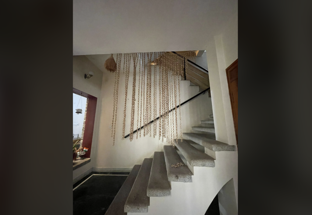 A modern interior staircase with concrete steps and metal railings ascends along a white wall. Hanging decorative beads cascade down the stairwell, adding an artistic touch. A window on the left allows natural light to brighten the area.