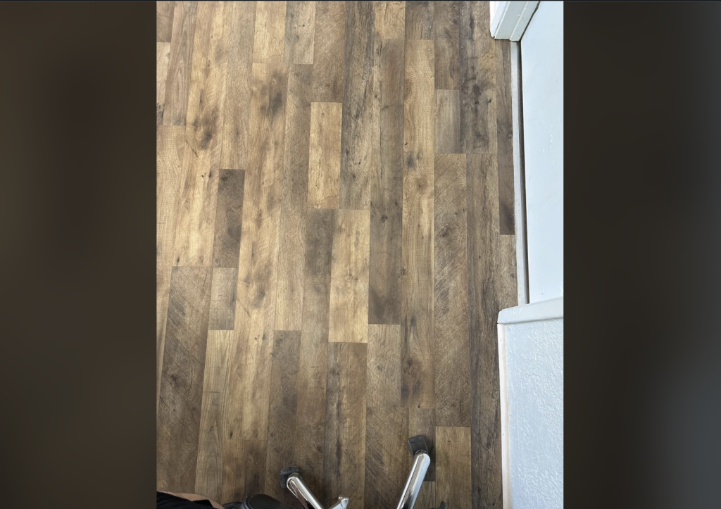 A wooden floor with varying shades of brown in a plank pattern is shown. The corner of a room with white walls can be seen. The bottom edge of a walker and a small part of a black shoe are visible at the bottom.