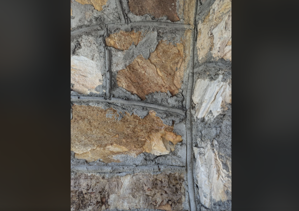 Close-up image of a textured stone wall with irregularly shaped, tan, brown, and off-white stones. The stones are set in gray mortar, which is visible between the stones, enhancing the wall's rough and rugged appearance.