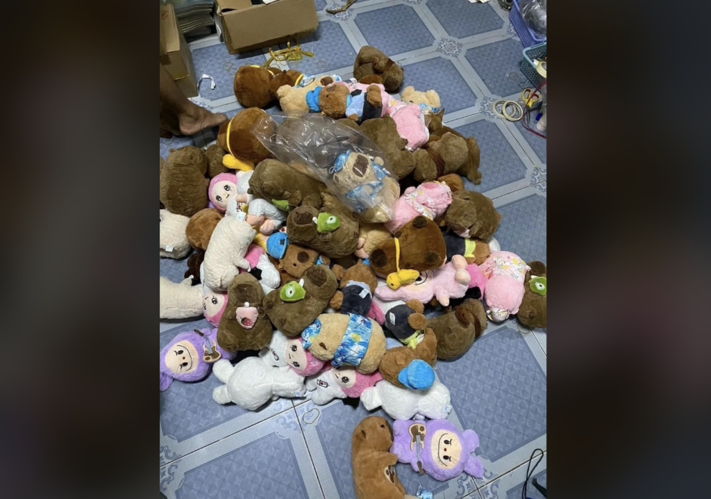 A large pile of plush toys, including bears, sheep, and other animals, is scattered on a tiled floor. Some of the toys are still wrapped in plastic. Various boxes and wires are visible in the background.