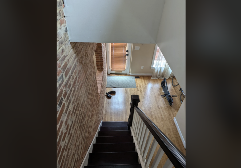 A view from the top of a staircase with dark wooden steps and a white railing. The stairway leads to a hallway with light wooden flooring, a brick wall on the left, and a bicycle leaning against the right wall. A pair of shoes is on a rug near the door.