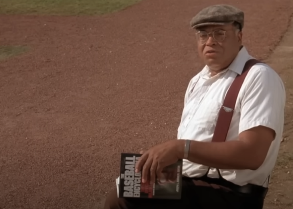 A man sits on a bench at a baseball field holding a book titled "BASEBALL PSYCHOLOGY." He is wearing a light shirt, suspenders, and a flat cap. The background features the infield of the baseball field and green grass.