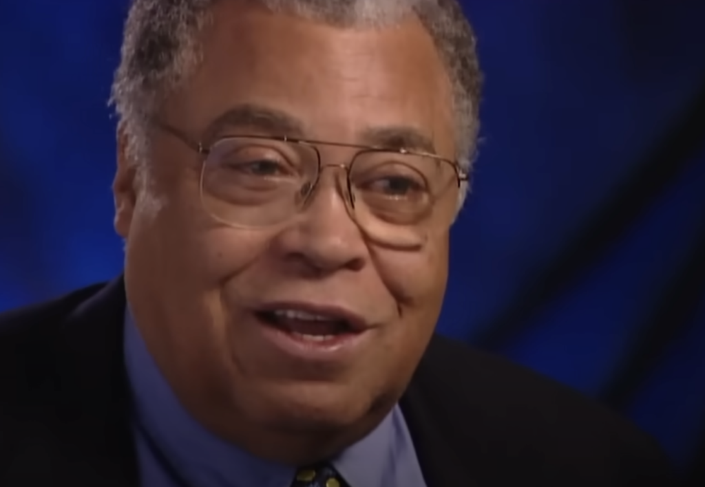 A person with short gray hair and glasses is smiling. They are wearing a dark suit jacket, a light blue shirt, and a patterned tie. The background is a gradient of dark blue.