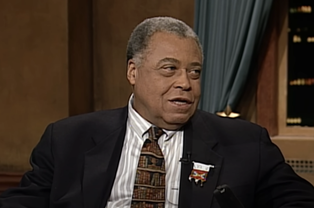 A person with short gray hair, wearing a dark suit and patterned tie, is sitting in a room with wooden paneling and green curtains. The person is looking to the side, smiling slightly.