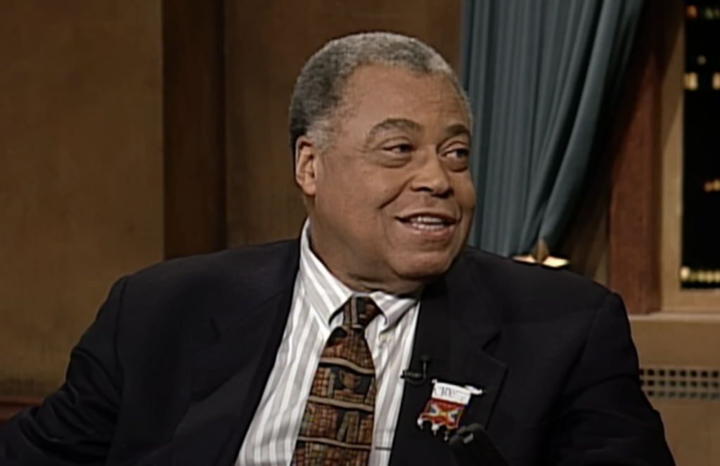 A smiling man in a dark suit and patterned tie is seated indoors. He is looking to his left. The background features brown walls, a curtain, and slightly blurred window lights.