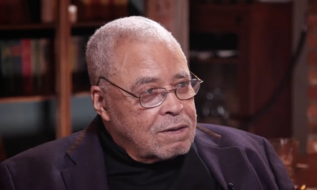 A man with gray hair and glasses is seated, wearing a dark suit jacket over a black shirt. He appears to be in a room with wooden shelves and cabinets in the background. His expression is calm and focused.