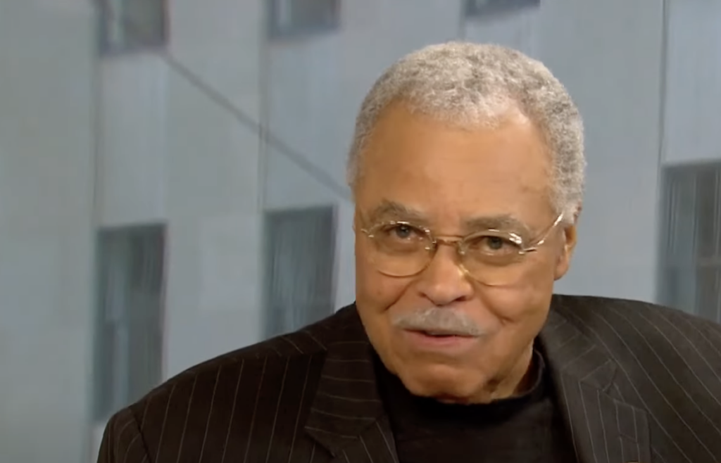 An elderly man with short gray hair and glasses is sitting indoors. He is wearing a dark pinstriped blazer and a black shirt. The background has large windows with blurred views of a building's exterior.