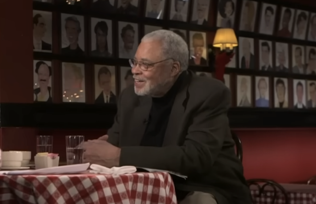 An elderly man with gray hair and a beard is sitting at a restaurant table covered with a checkered tablecloth. The table has glasses and a cup. The background features framed pictures and a lamp on the wall. He appears to be in conversation, smiling.