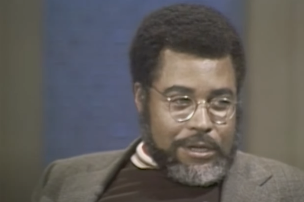 A man with glasses and a beard is seated against a blue background. He is wearing a dark blazer over a turtleneck sweater. The image appears to be from a television interview or talk show.