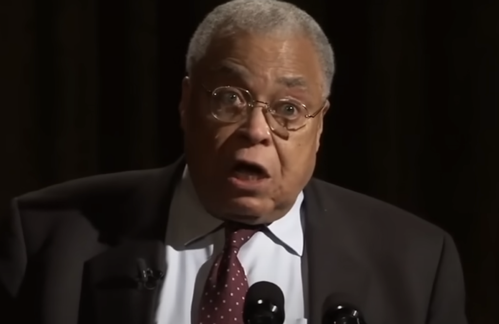 An elderly man with gray hair and glasses is speaking into a microphone. He is wearing a dark suit, a white shirt, and a red tie. The background is dark, and his expression appears serious.