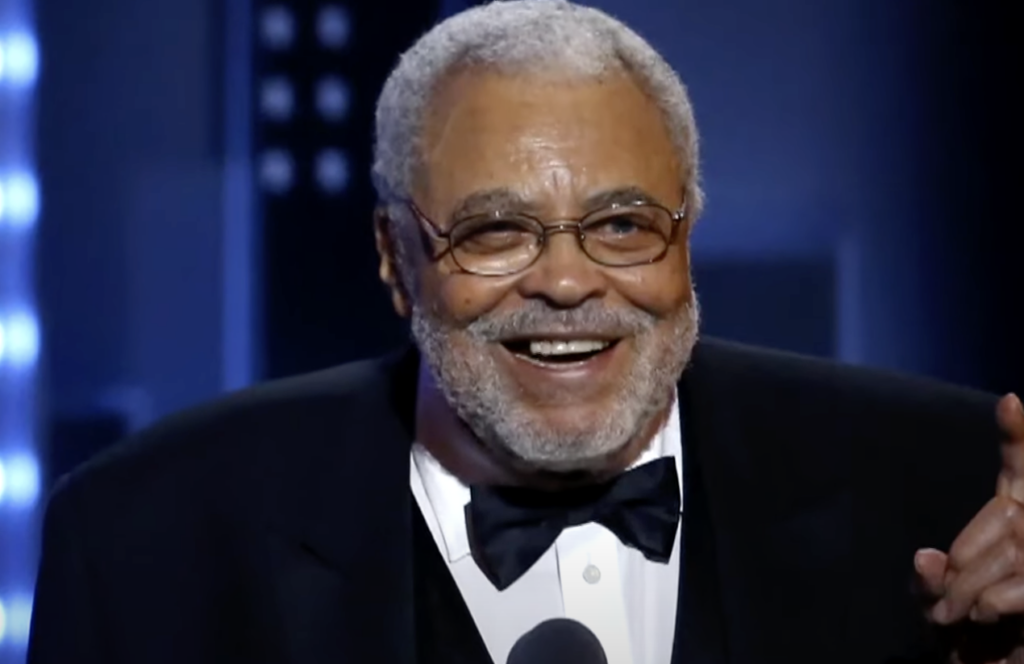 An elderly man with gray hair and a beard is wearing glasses, a black tuxedo, and a black bow tie. He is smiling and speaking into a microphone on a stage with an unfocused background of lights.