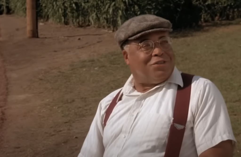 A man wearing glasses, a flat cap, white shirt, and brown suspenders smiles while sitting outdoors on a grassy area. There’s a dirt path and tall greenery in the background. The scene appears to be rustic and set in a rural area.