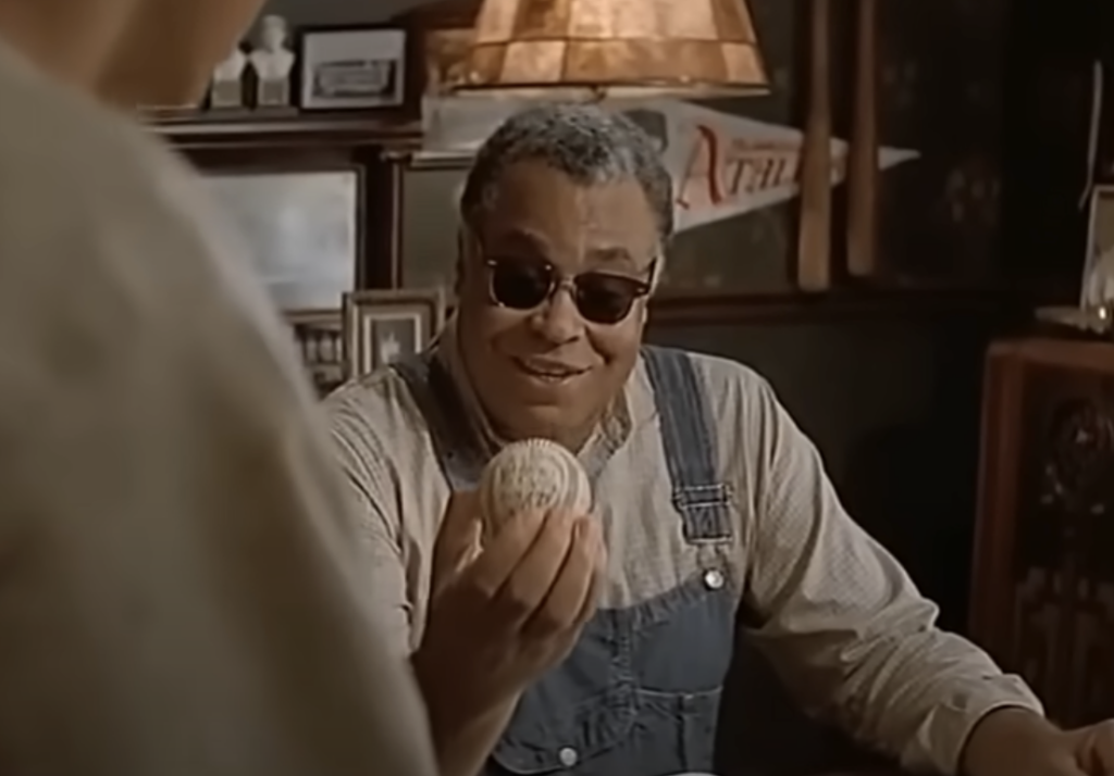 A man with gray hair and sunglasses, wearing blue overalls and a beige shirt, holds a baseball while looking at another person. The dimly lit room has a cozy decor with wooden furniture and warm lighting. A pennant and framed pictures adorn the background.