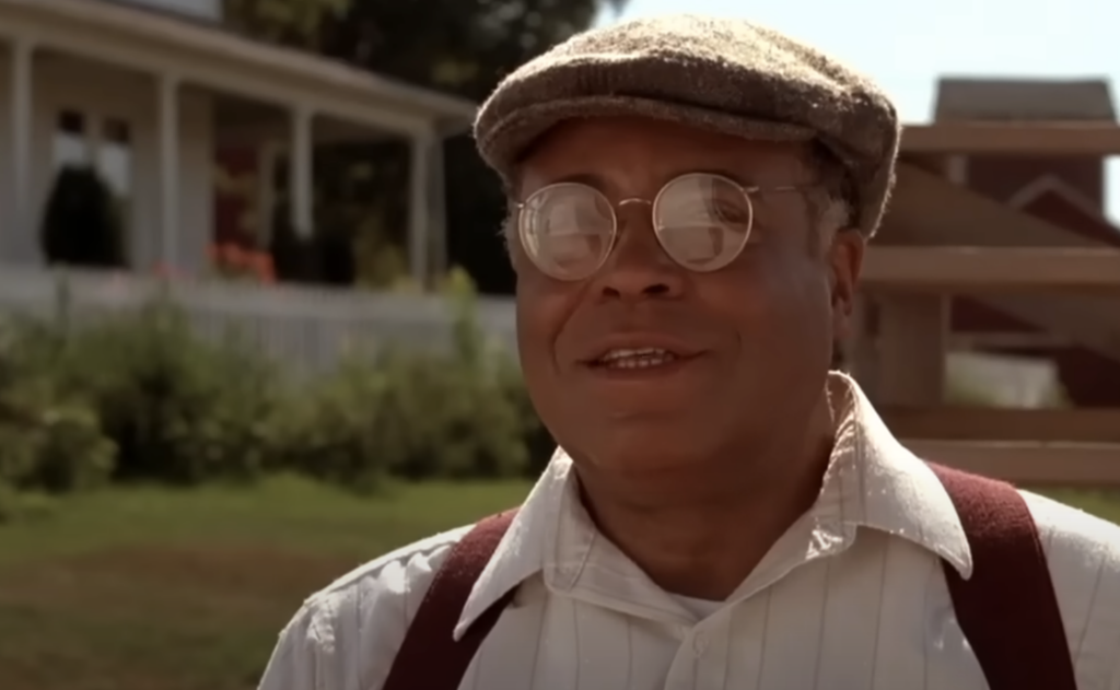 An older man wearing round glasses, a flat cap, a white shirt, and brown suspenders stands outdoors in front of a house with a porch. He is smiling slightly and the sun is shining brightly. The background includes greenery and parts of the house.