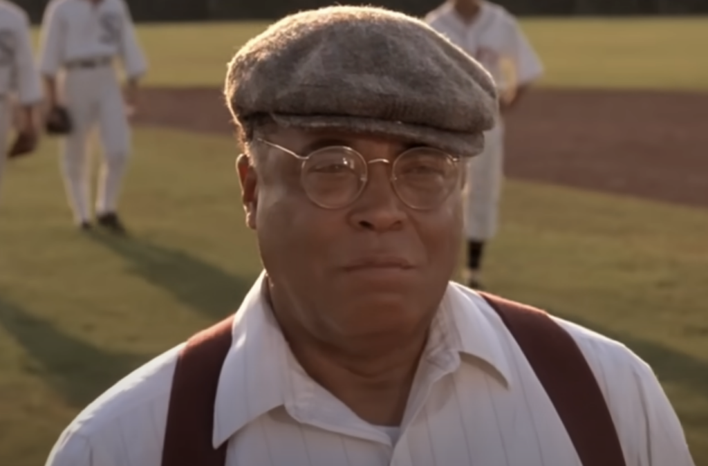 A man wearing a cap, glasses, white shirt, and suspenders stands on a baseball field. Several players in white baseball uniforms are visible in the background. The man has a determined expression on his face. The setting appears to be a sunny day.