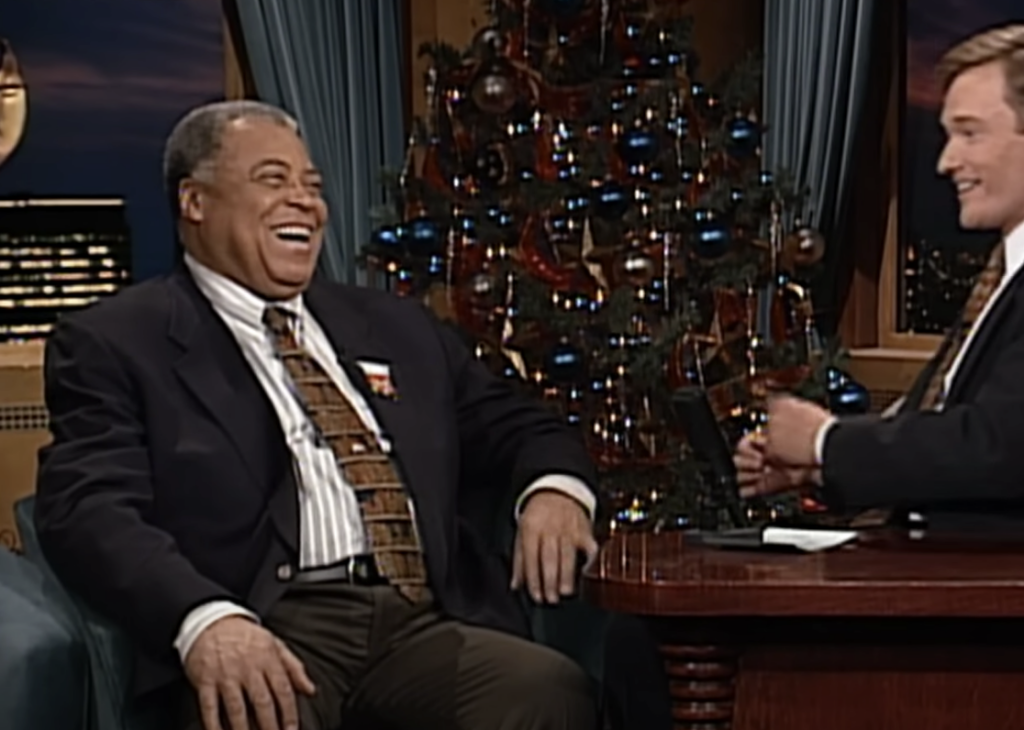 Two men are seated in a talk show setting, smiling and engaged in conversation. Behind them is a decorated Christmas tree. One man is wearing a dark suit jacket with a patterned tie, and the other is also in a suit, seated at a desk with a microphone.