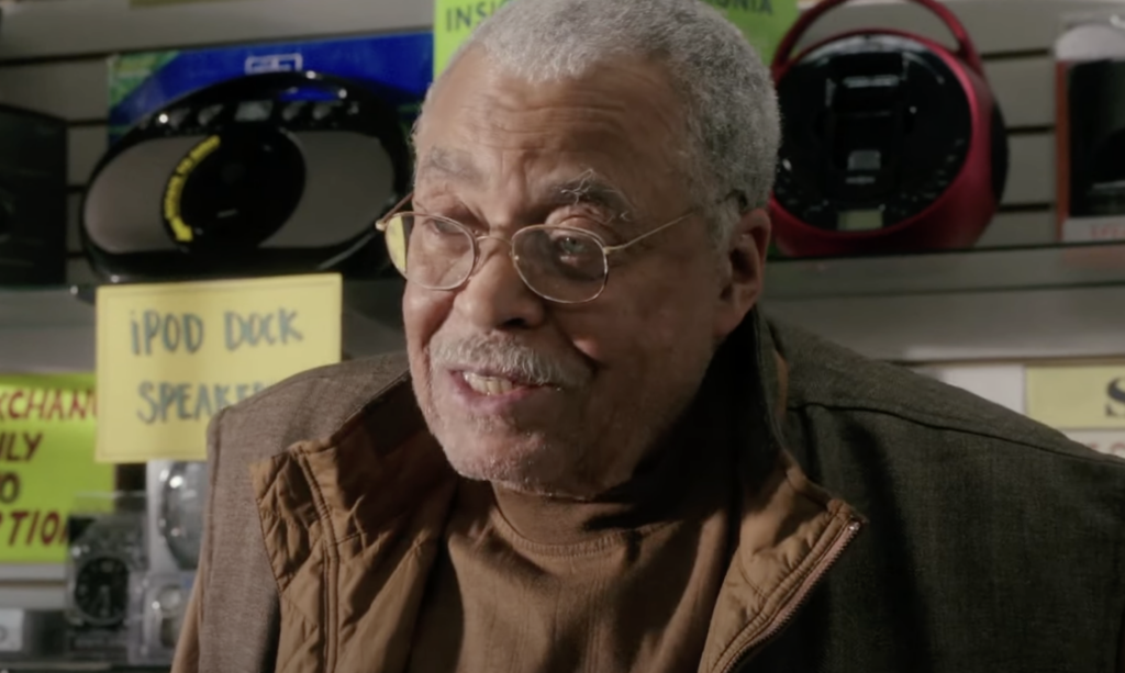 An elderly man with glasses and a mustache is seen smiling inside a store. He is wearing a brown jacket and stands in front of shelves displaying various electronic items, including an iPod dock and speakers with visible price labels.