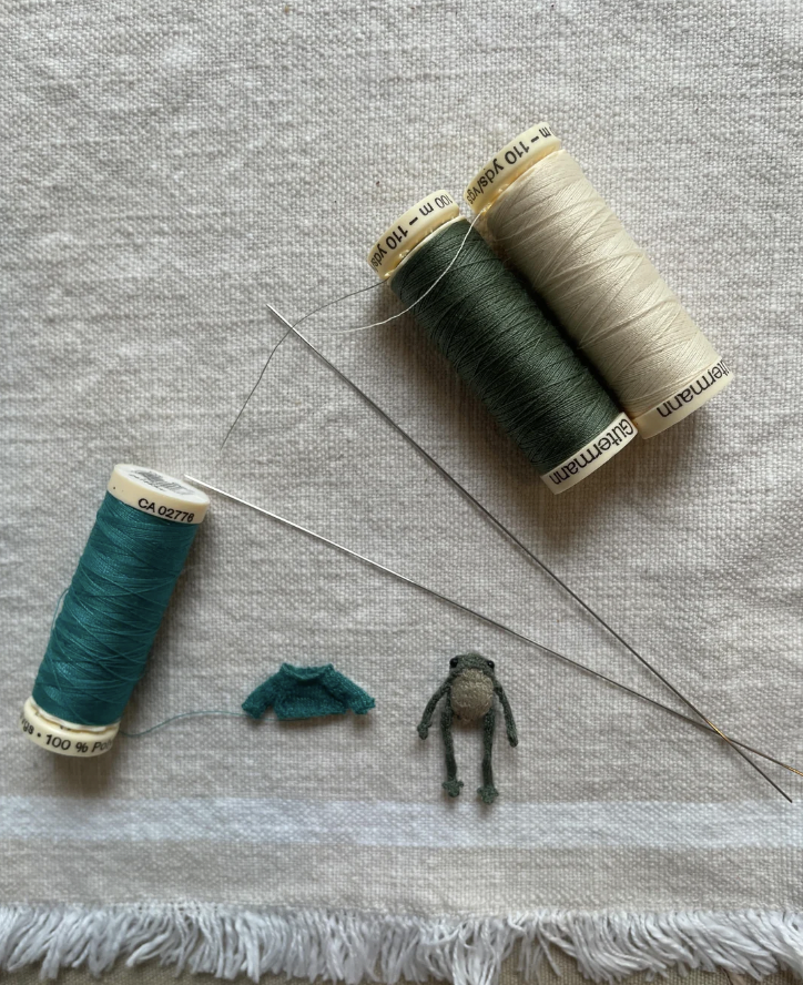 Three spools of thread in teal, olive green, and beige lie on a fabric surface with a needle next to them. Below, there is a tiny handmade frog figurine and a miniature knitted sweater, all crafted with the same colored threads.